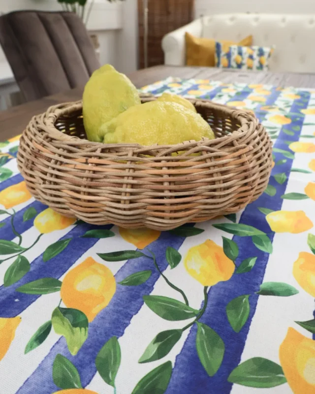Zitronen Tischläufer "Lemontree" auf Holztisch im Esszimmer von Sander. Der Tischläufer ist blau weiß gestreift mit vielen kleinen Zitronen als Motiv. Auf dem Tischläufer steht eine Rattan Schale mit 3 Zitronen. Im Hintergrund sind 1 Stuhl und ein Sofa erkennbar.