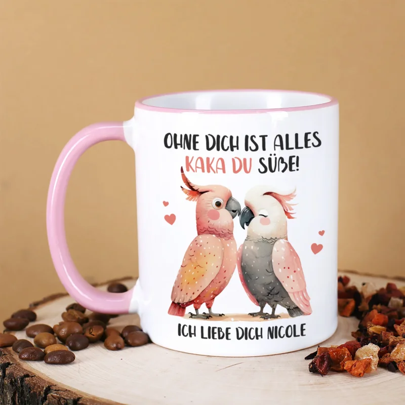 Tasse mit Kakadu-Motiv und Spruch „Kaka du Süße!“ von der Seite, rosa Henkel sichtbar, dekoriert mit Kaffee und Tee